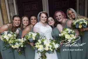 bride wears white dress, red hair, ginger hair, white and green flowers, flora arrangement, six bridesmaids, surround bride, smiling, close up