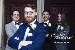 groom in foreground, beard, spectacles, arms folded, navy suit, white button hole, smiling groomsmen behind, wearing different attire