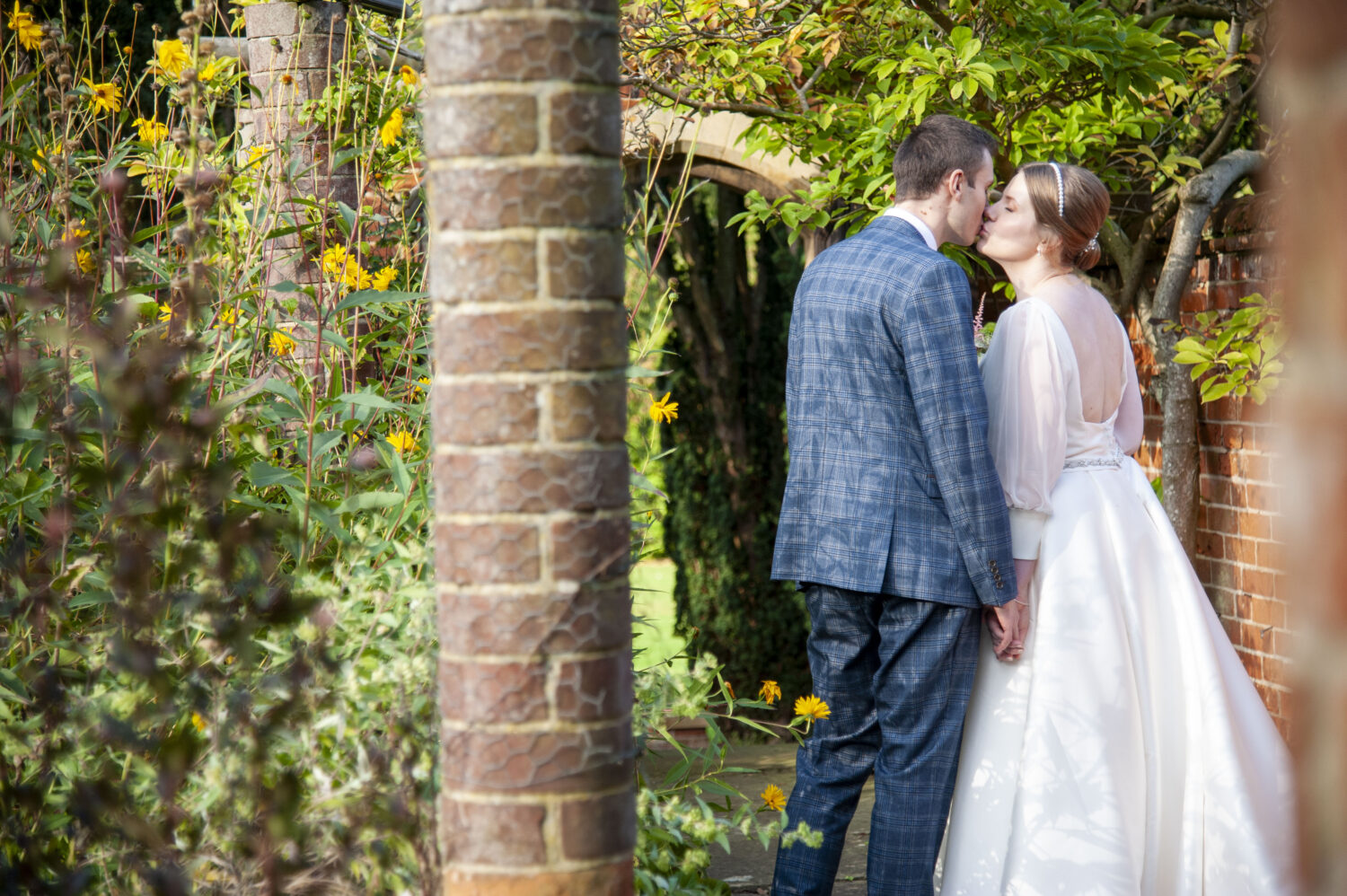 lanwadeshall, bride groom kissing, holding hands, garden, blue check suit, white dress,brick column,