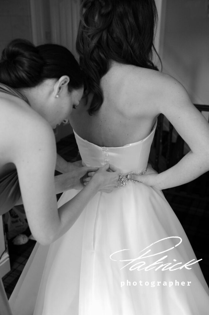 bridal preparation, white satin dress, buttons, beaded and glass belt. Posterior shot. Bride long dark hair. Bride hands on waist. Bridesmaid, dark hair tied chignon. Doing up wedding dress