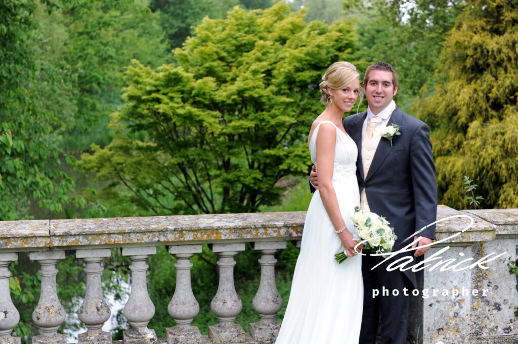 braxted park bridge river trees bride carries bouquet, groom has hand on her back