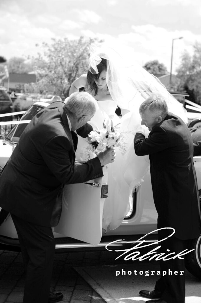 black and white image. bride climbs out of car. helped by dad and chauffeur. Chaffeur holds bouquet. White veil blows in the veil