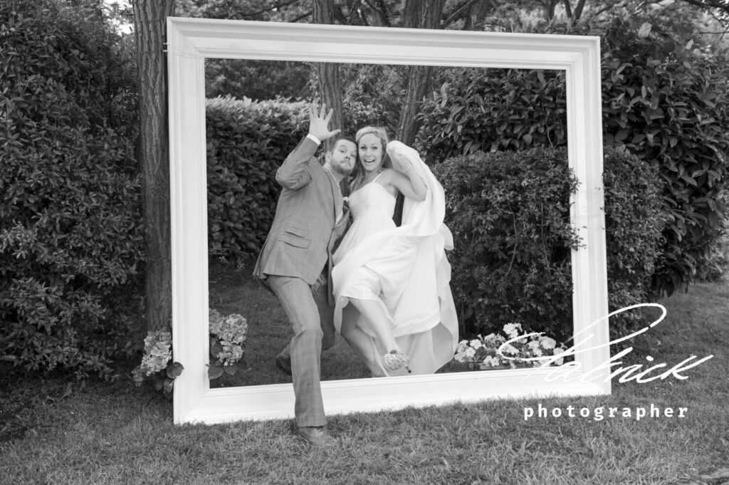 black and white image. Bride lifts dress. Playful image bride and groom, giant frame in garden setting