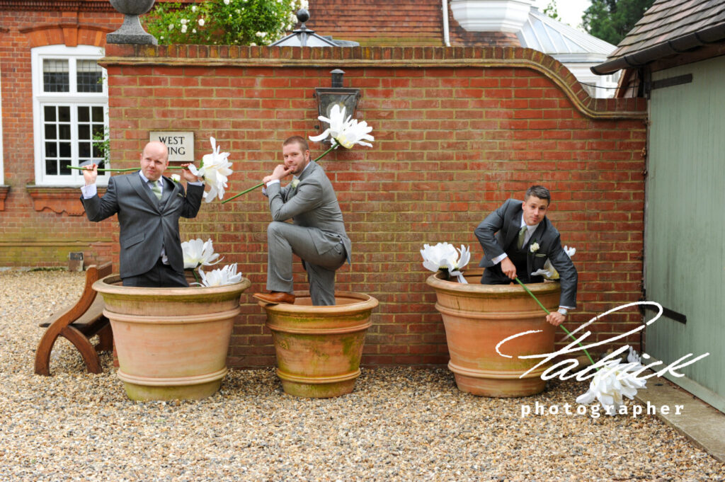 flowerpot men. Playful groomsmen, grey suits, giant flowers in giant flowerpot. Brick wall, house in background, gravel drive