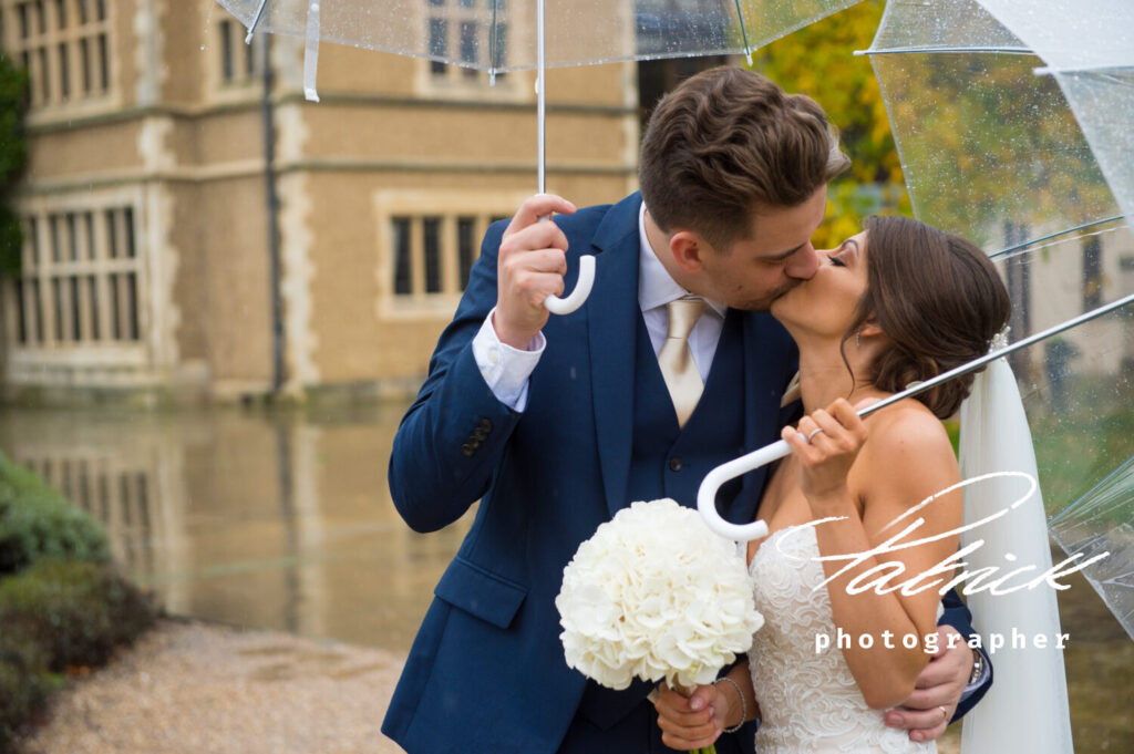 close up bride and groom kissing under umbrellas in the rain, blue suit pale gold tie, white dress, white bouquet