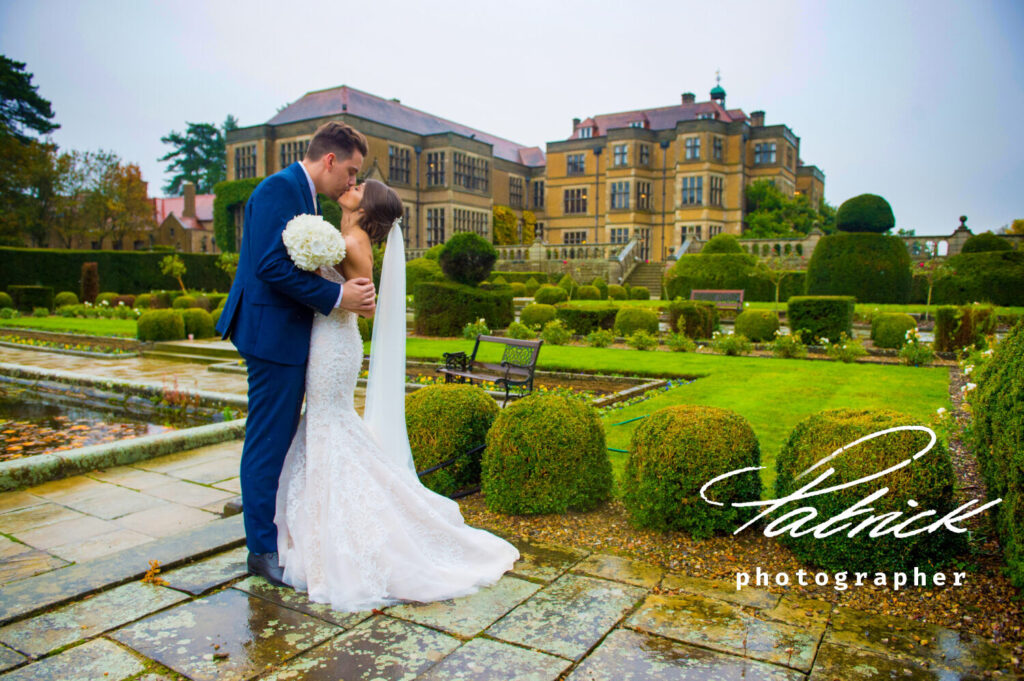 wet wedding day, fanhams hall background, bride and groom kissing foreground. white bridal grown, white bouquet and blue suit, daytime image