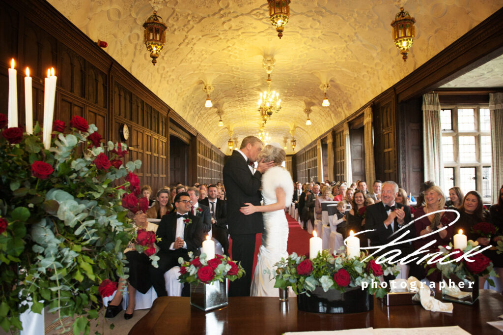 fanhams hall, wedding ceremony, winter wedding, candlelit, wedding guests, red carpet, red and white floral, bride and groom kissing, romantic, onlooker sitting, clapping, celebration