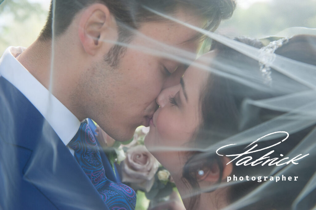 bride and groom kissing under veil. royal blue jacket and blue and purple tie, bride wears crystal and pearl tiara, pearl earrings, pale pink rose buttonhole