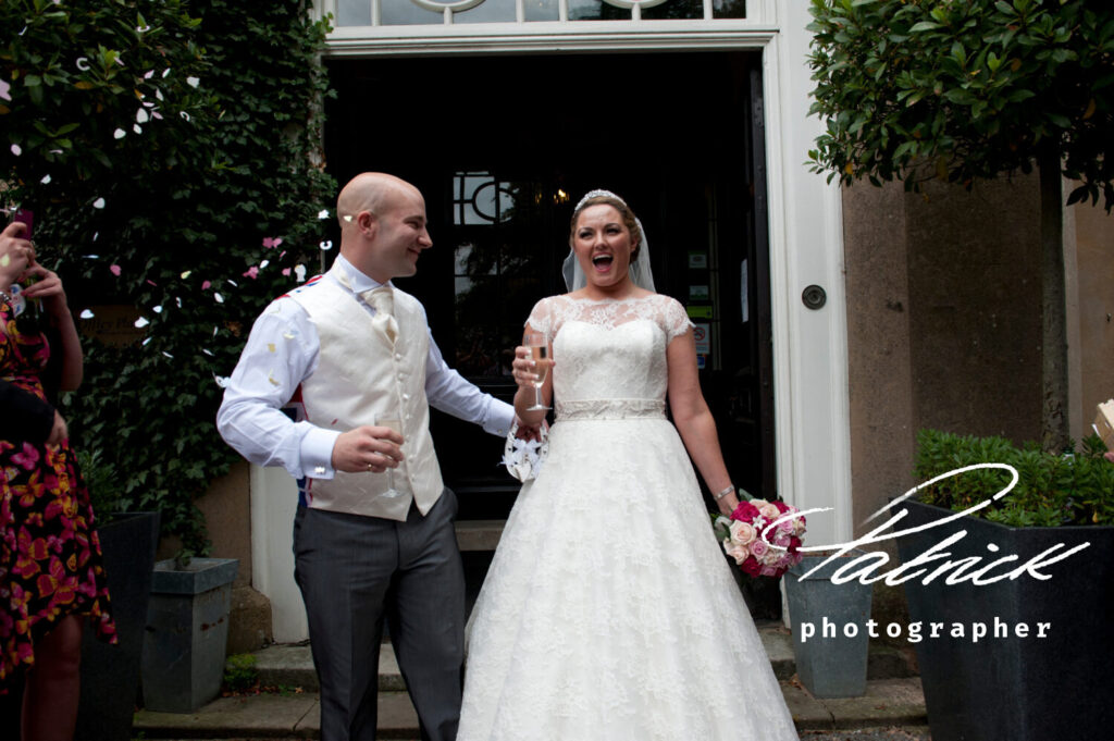 bride exclaiming whilst groom looks on. bride carries champagne and prink and white bouquet