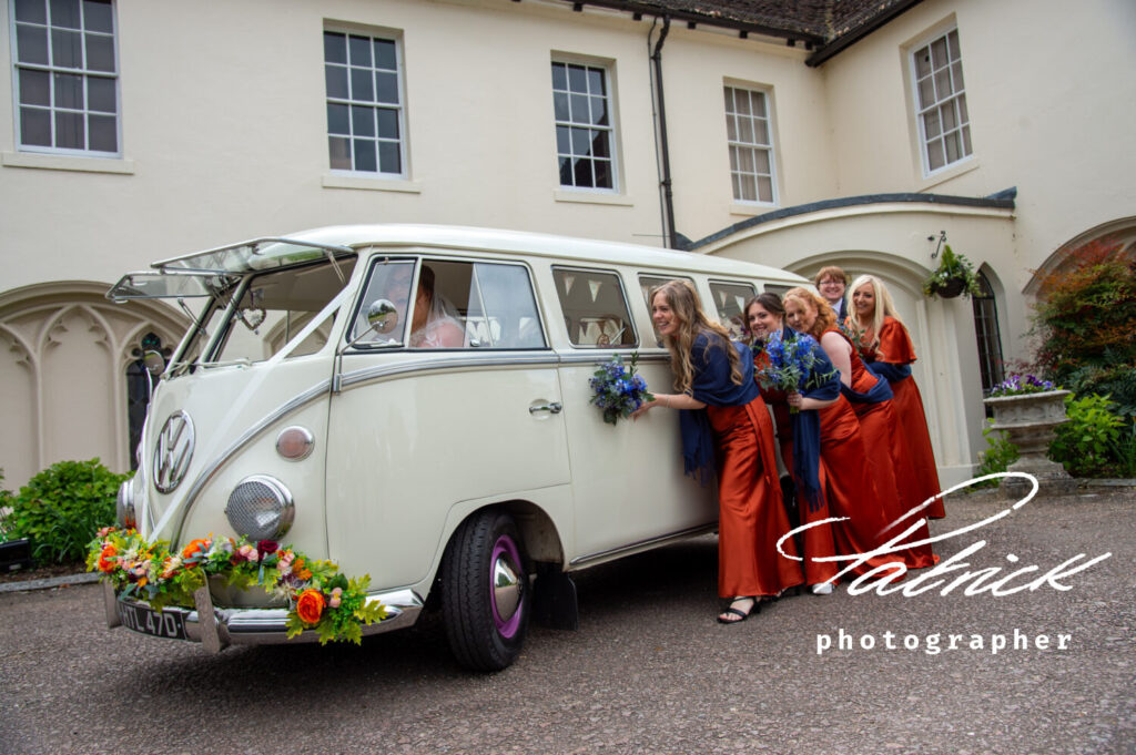 lucky penny camper van, decorated with flowers, bridesmaids pretend to push from side. orange dresses, navy shawls and bouquet, ware priory exterior,