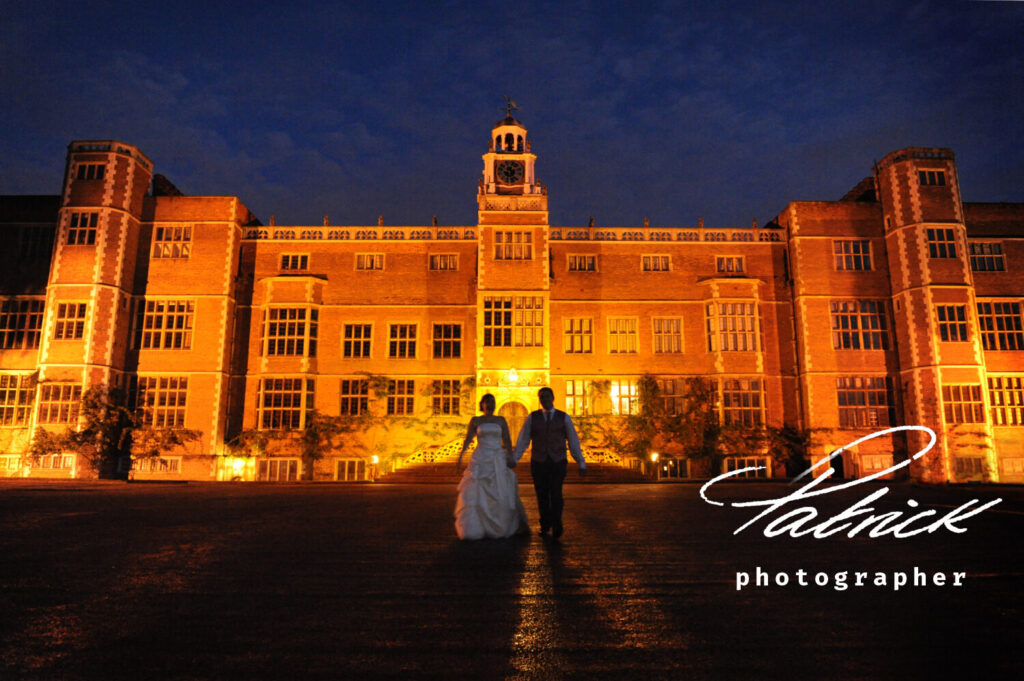 in the background illuminated Hatfield House, exterior, at night, dark sky. Bride and Groom walk towards camera