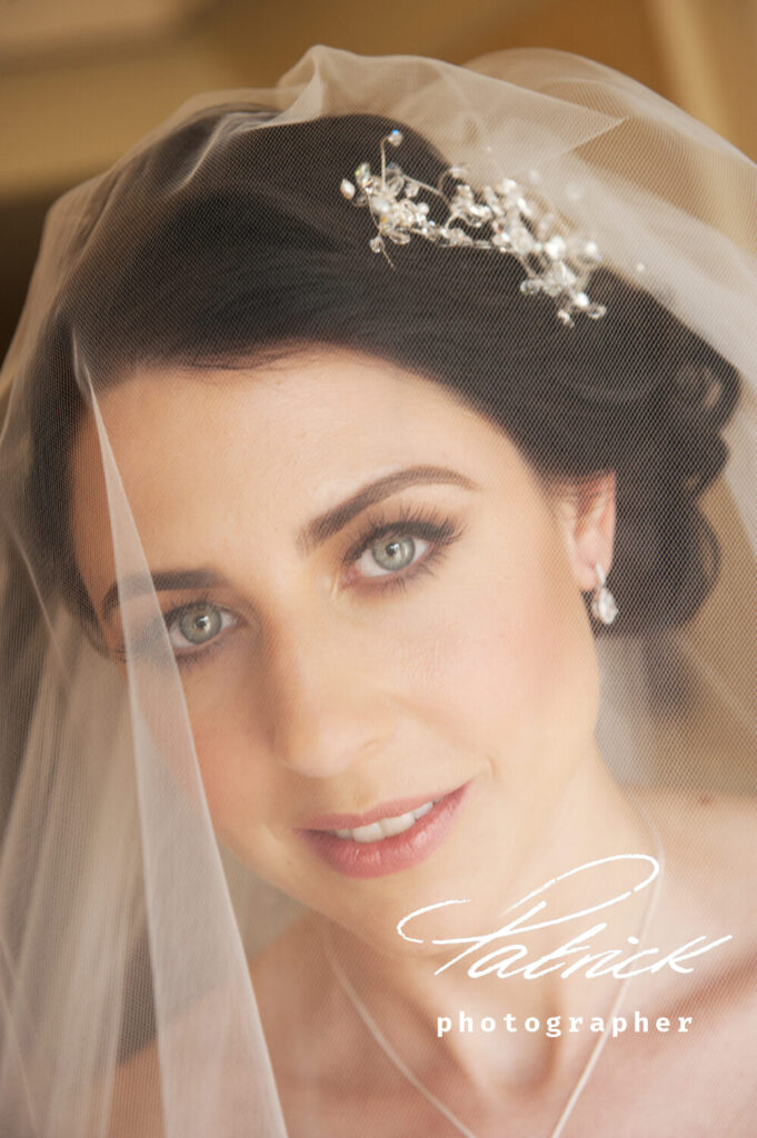 bride looking at camera, under the veil. Dark hair, green eyes, wearing crystal and pearl hair accessories and crystal earrings.