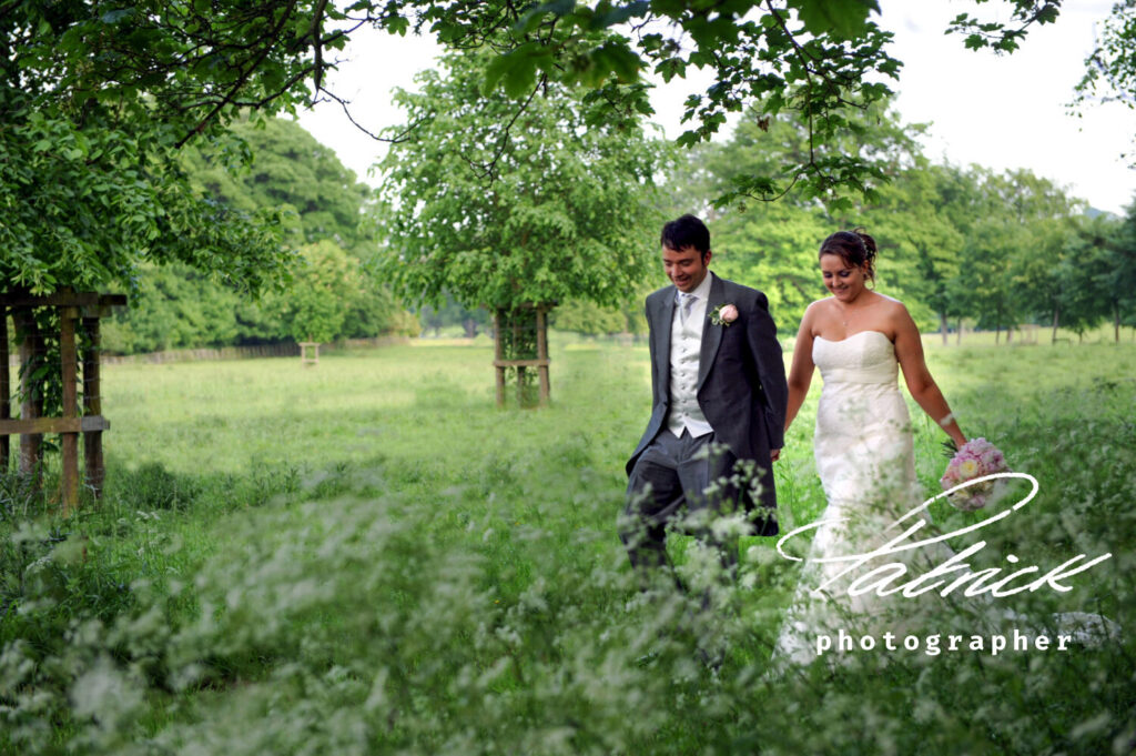 bride and groom walking through grounds at Hatfield House, green trees, long grass, pink bouquet