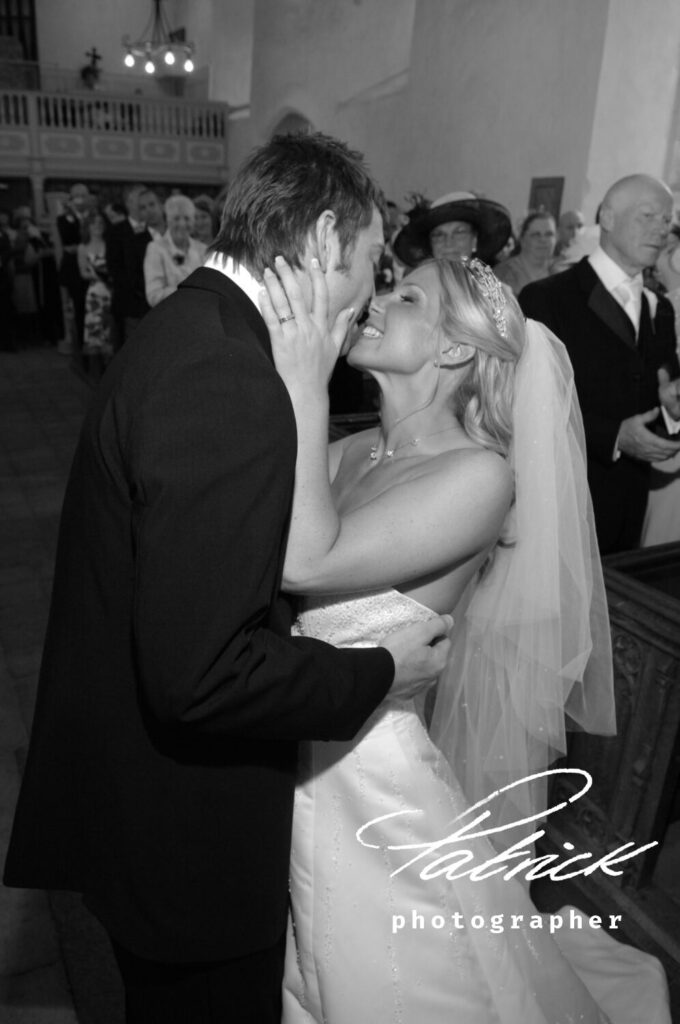 black and white image, bride clasps grooms face in her hands. wedding guests look on. at alter inside church