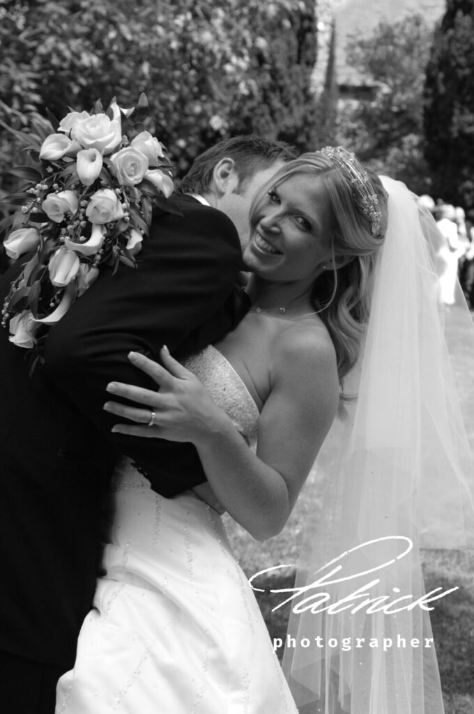 bride smiling at camera, groom embraces her, bouquet, black and white image