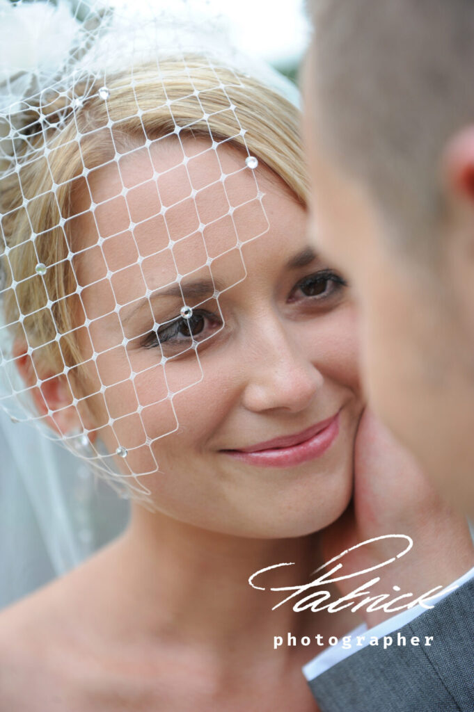 groom holds brides face in right hand, she looks at him . white net fascinator