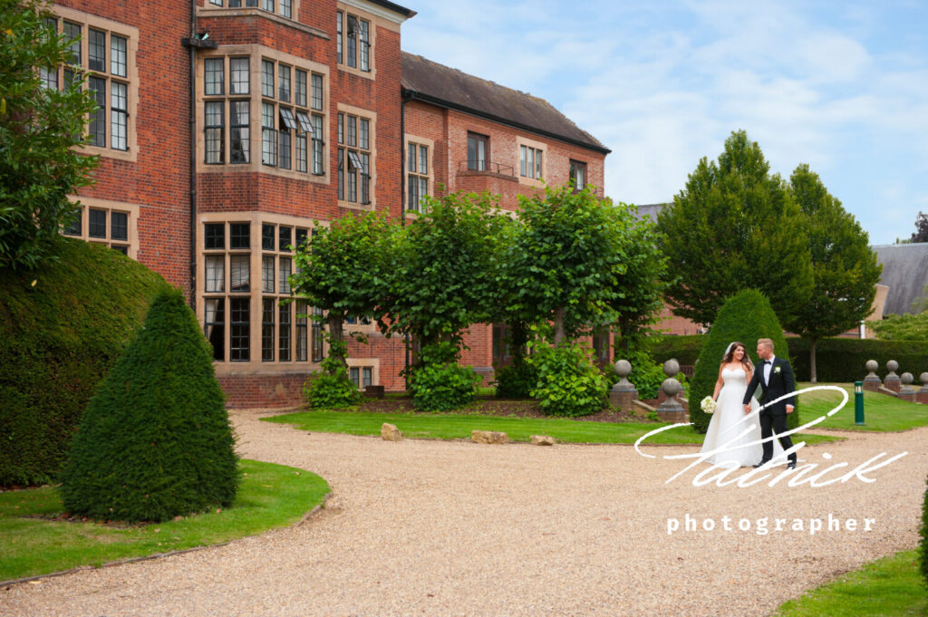 Bride and groom walk across the gravel drive in front of the exterior of Hanbury Manor in Ware.©patrickphotographer.com