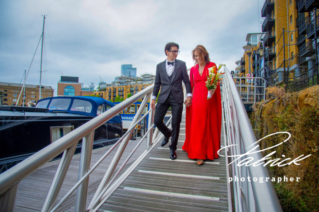 st katharine docks London bride wears long red floaty dress carries bouqet of lilies Holds her new husband's hand as they walk down to the boat