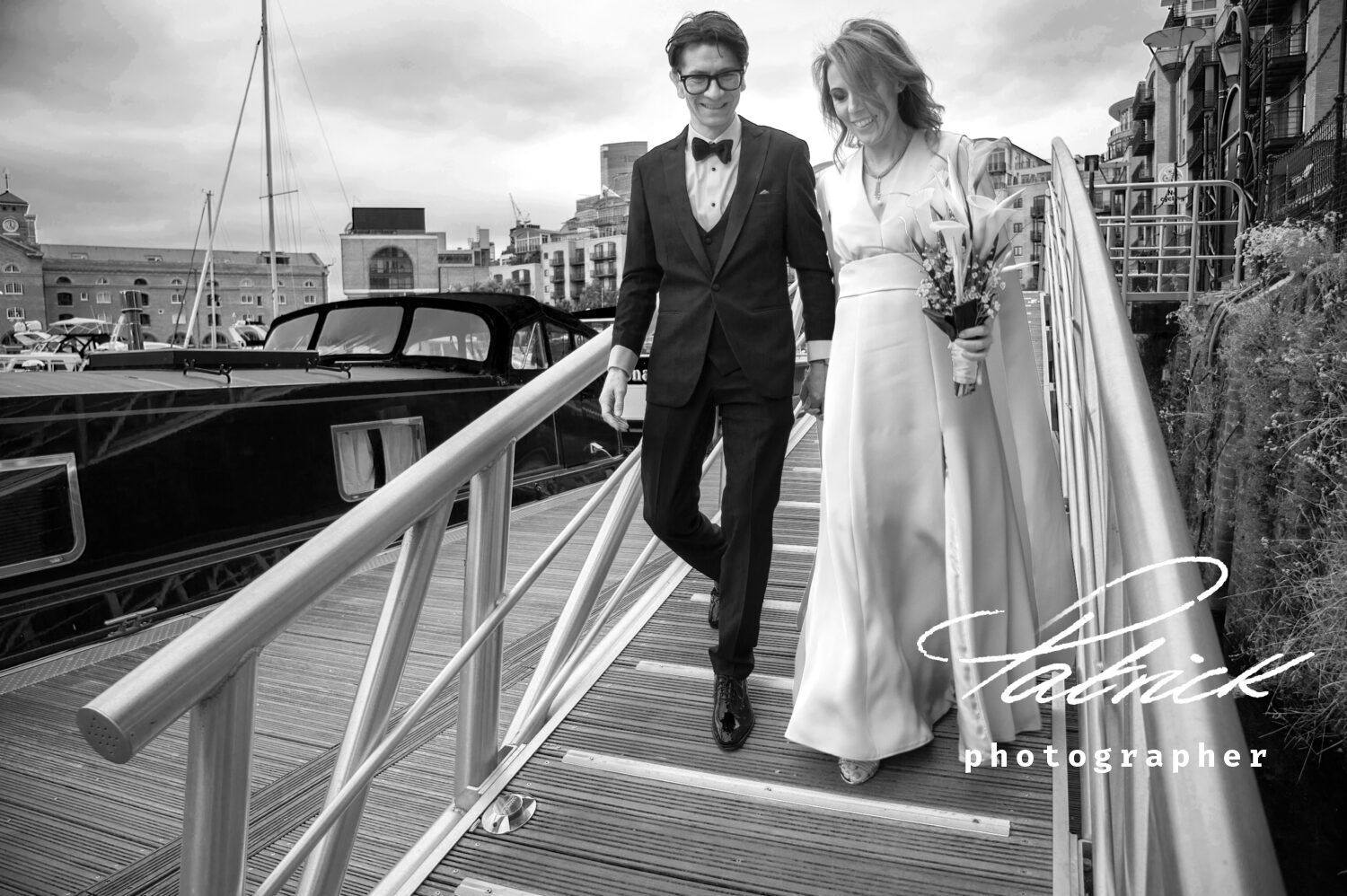 black and white image of bride and groom walking down to st katharine dock London boats and London in the background