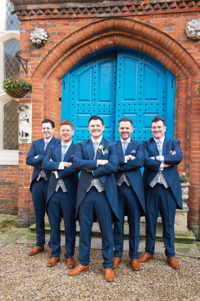 exterior gosfield hall, blue door, groom and groomsmen smiling, looking at camera, arms crossed