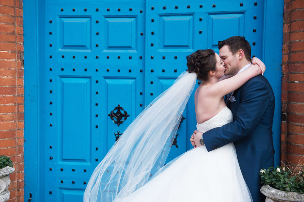 bride embraces her new husband, arms wraps around his shoulders, his around her wait. big embrace and kiss. veil blows in wind. exterior of blue door, gosfield hall
