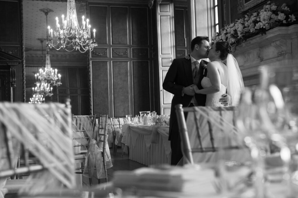 Grand Ballroom and Queen Charlotte’s Oak Panelled Gallery. bride and groom kiss, long windows and chandeliers, tables dressed for wedding breakfast, black and white image