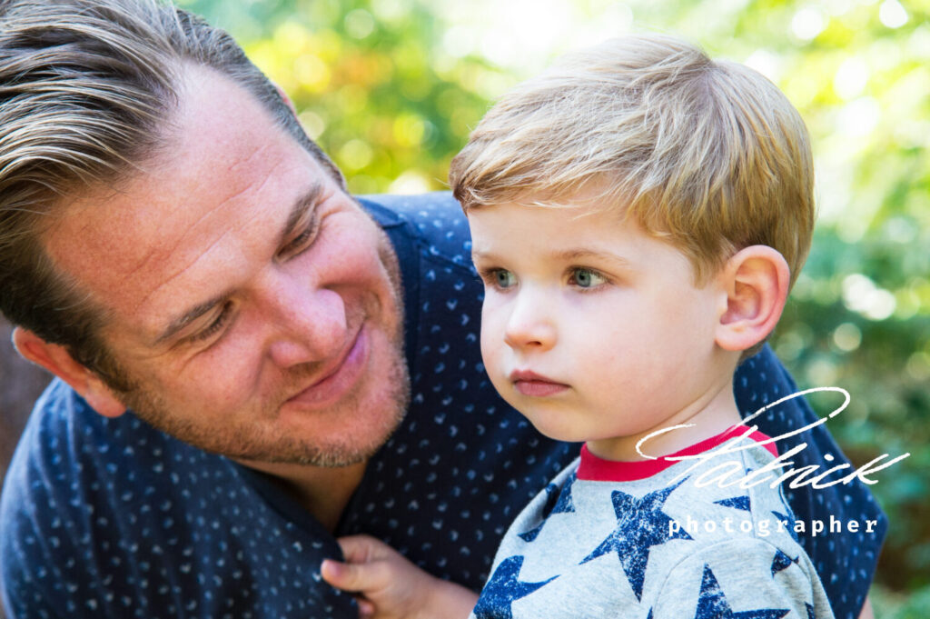 lifestyle, portrait, father and son. boy is five, dad wears blue shirt, has swept back hair. Dad smiling at son. Son looking forward and is distracted. Fair haired oy wears t-shirt with blue stars.